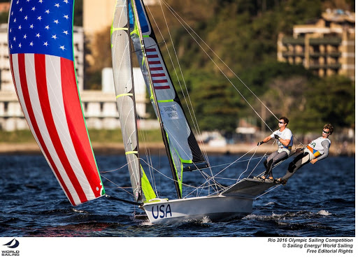 Two athletes compete in a sailing event at the Rio 2016 Olympics, steering a boat with a large, colorful sail featuring the American flag. The boat cuts through the water with buildings and trees in the background.