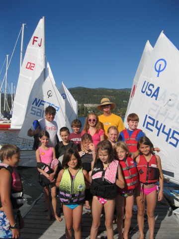 A group of children and two adults wearing life jackets stand on a dock in front of sailboats on a sunny day, with mountains and a clear blue sky in the background.