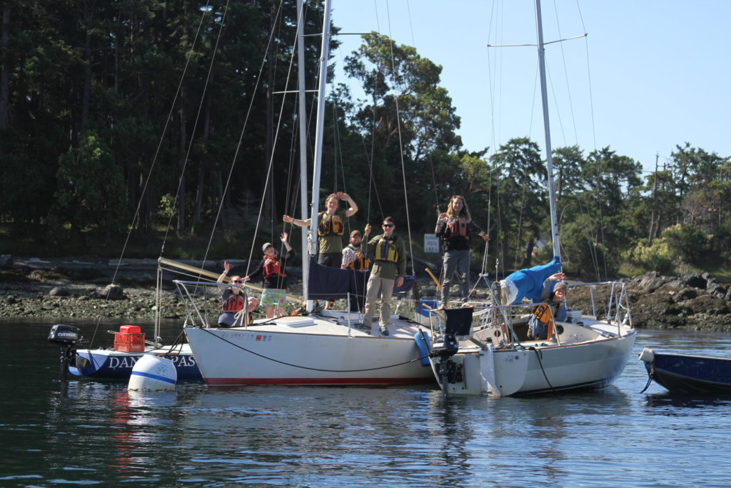 A group of people wearing life jackets stand and sit on two docked sailboats in calm water, surrounded by trees and greenery on a sunny day. Some are looking through binoculars or waving.