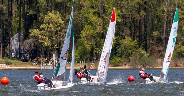 Several small sailboats with teams of people race on a lake, passing red buoys, with trees and a house visible in the background on a sunny day.