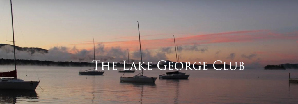 Sailboats anchored on a calm lake at sunrise or sunset, with mist rising from the water and 'The Lake George Club' text overlayed in white.