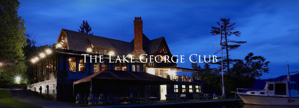 A large lakeside lodge with warm lights and a brick chimney sits beside a docked boat at dusk. Text over the image reads 'The Lake George Club.