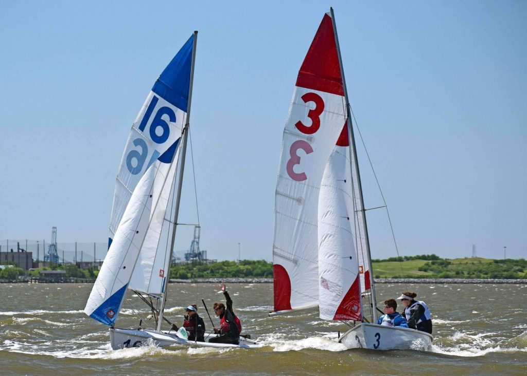 Two small sailboats with white sails, marked with blue "16" and red "3", race on choppy water under a clear sky. Teams of sailors in life jackets are actively steering and adjusting the sails. Industrial structures are visible in the background.
