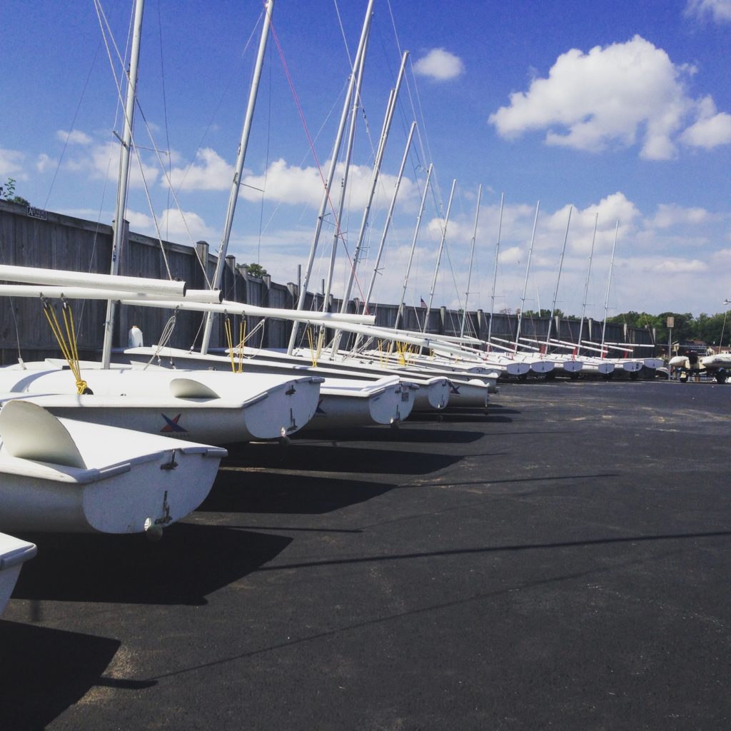 A row of white sailboats with tall masts are lined up on an asphalt surface under a sunny blue sky with a few clouds, next to a tall concrete wall.