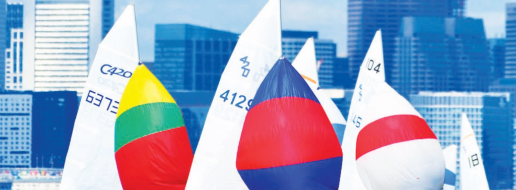 Colorful sailboats with bright spinnakers and tall white sails race in front of a city skyline, with modern high-rise buildings visible in the background under a clear blue sky.