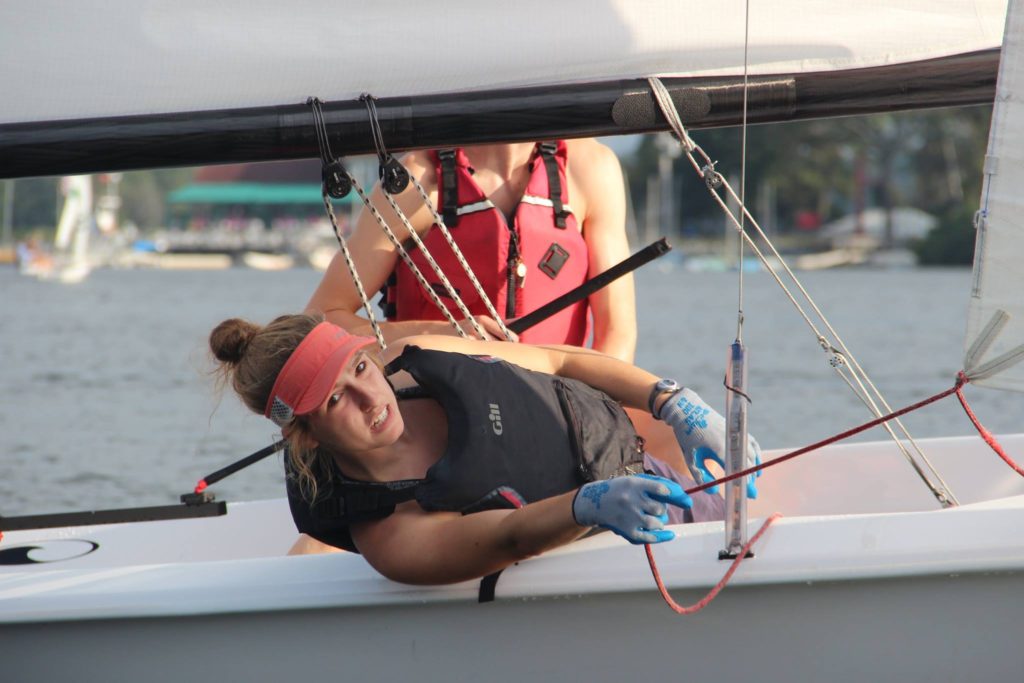 A person wearing a red visor, blue gloves, and a life jacket leans out of a sailboat, adjusting a line while another person in a red vest stands behind. The boat is on a lake with other boats and trees in the background.