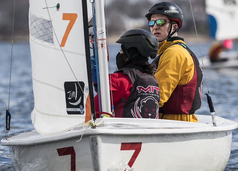 Two people in helmets and life jackets sail a small boat marked with the number 7 on the water. The person in front wears an "MIT Engineers" vest; the other is in yellow and looks back, both focused on sailing.
