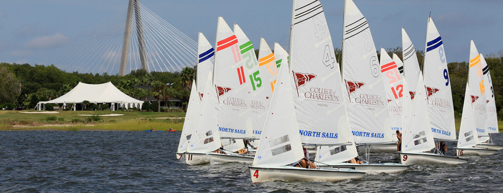 Several small sailboats with colorful numbers and stripes are racing on the water near a grassy shoreline, with a white tent and a large cable-stayed bridge visible in the background.