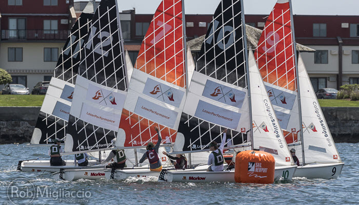 Four small sailboats with colorful sails race around an orange buoy on a sunny day, with sailors focused and buildings visible in the background. The sails display sponsor logos and numbers.