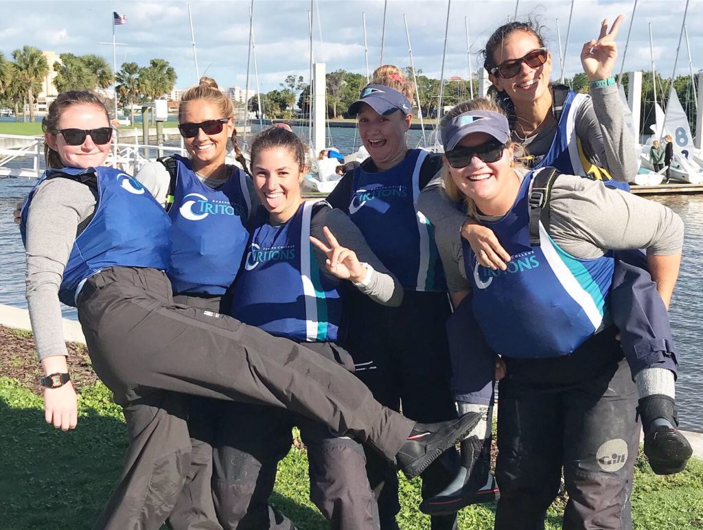 Six women in matching blue CURCIONS vests and outdoor gear pose cheerfully by a marina, some making peace signs; boats, palm trees, and a dock are visible in the background.