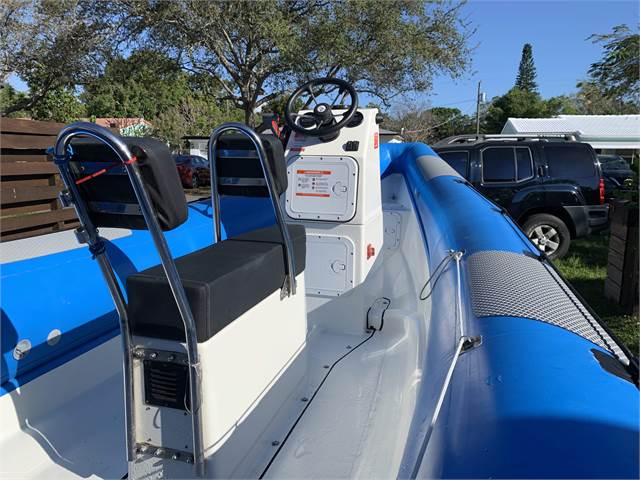 A blue and white inflatable boat with steering console and black bench seats is parked on grass. Trees, cars, and houses are visible in the background on a sunny day.