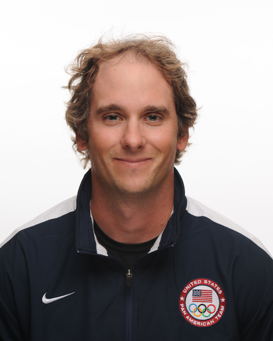 A man with light brown curly hair wearing a dark jacket with the United States Olympic Team logo and a Nike logo, smiling in front of a plain white background.