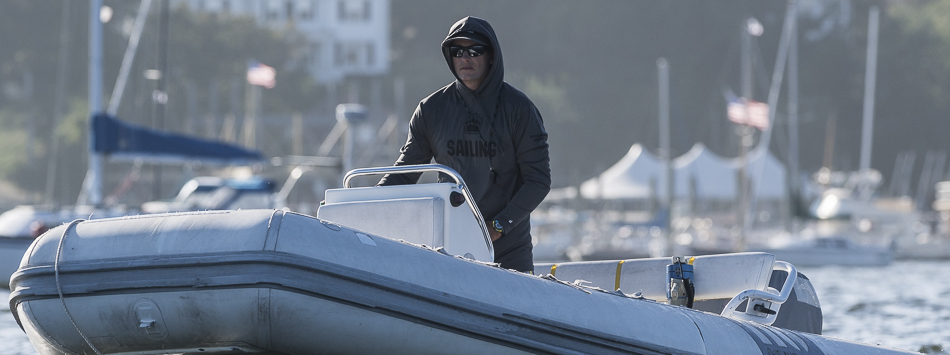 A person wearing sunglasses and a hooded jacket stands at the controls of a gray inflatable boat on the water, with sailboats and American flags visible in the blurry background.