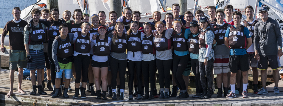 A group of around 30 young adults in matching “Brown” athletic shirts stand smiling together on a dock in front of sailboats, posing for a team photo in sunny weather.