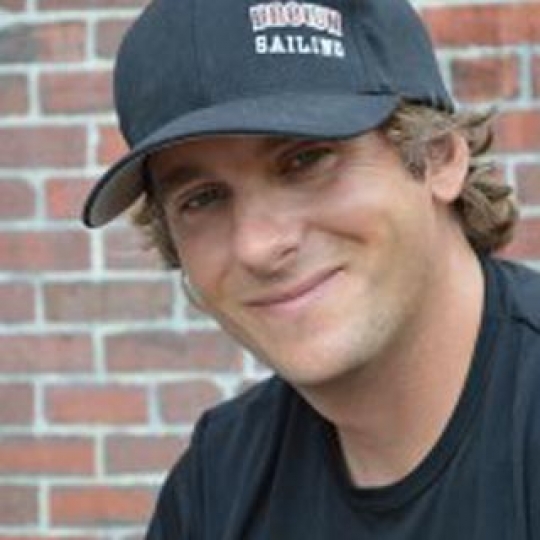 A smiling man wearing a black baseball cap and black T-shirt stands in front of a brick wall. The cap has white and red text that reads 'Brown Sailing.