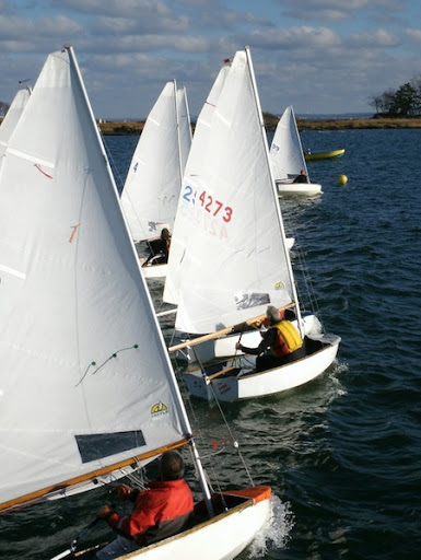 Several small sailboats with single sailors compete in a race on a sunny day, moving through blue water towards a yellow buoy under a partly cloudy sky. The sailors are dressed in life vests.