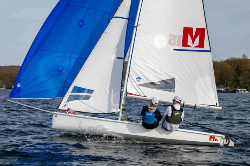 Two people in life jackets sail a small white sailboat with blue and white sails on a lake, with trees and shoreline visible in the background.