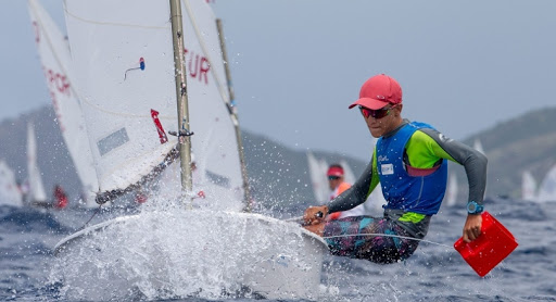 A sailor in bright clothing and a red cap leans out of a small sailboat, splashing water as they navigate choppy waves during a race. Other sailboats are visible in the background.