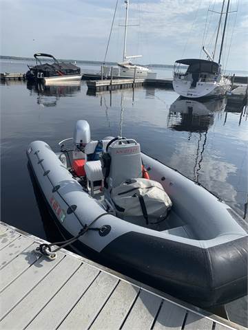An inflatable gray motorboat is docked at a marina with other boats and yachts moored nearby. The boat has an outboard engine and equipment inside, and calm water reflects the vessels and sky.