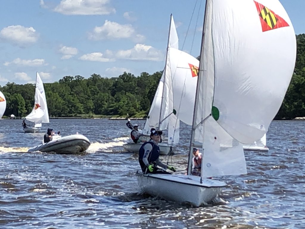 Several small sailboats with white sails and colorful emblems race on a lake, with a motorboat in the background and tree-lined shore under a blue sky with scattered clouds.