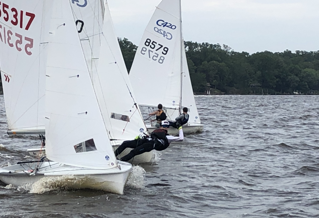 Three small sailboats with sailors leaning out over the water race closely together on a choppy lake, with trees lining the distant shore under a cloudy sky.