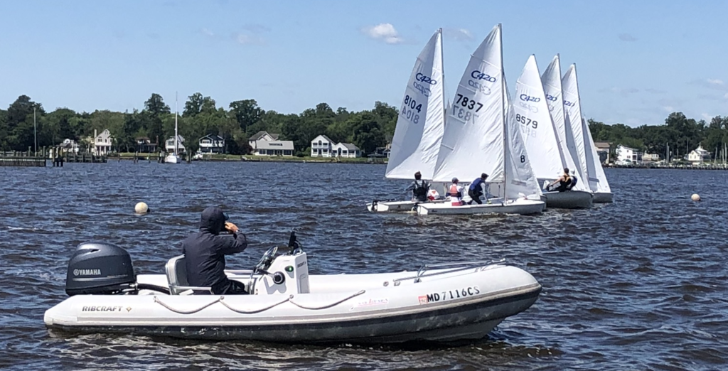 A person in a motorized inflatable boat observes several small sailboats with white sails racing on a river near shore, with houses and trees in the background under a blue sky.