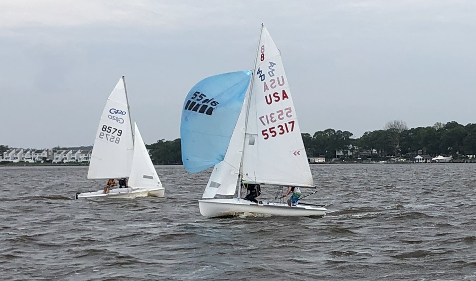 Two small sailboats race on choppy water near a shoreline with trees and houses. The boat in front has a blue spinnaker sail and the numbers "55317 USA" on its mainsail. The second boat sails slightly behind.