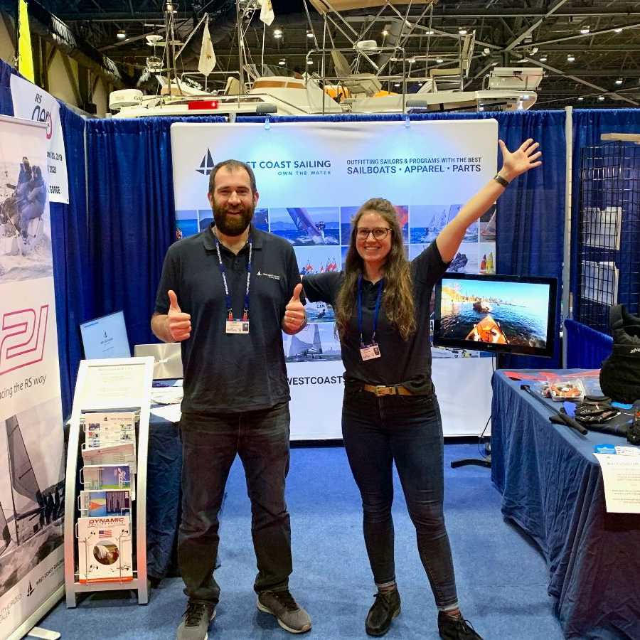 Two people stand smiling in front of a West Coast Sailing booth at an indoor event. The man stands with his arms at his sides, and the woman raises one arm cheerfully. Sailing gear, signs, and display monitors are visible.
