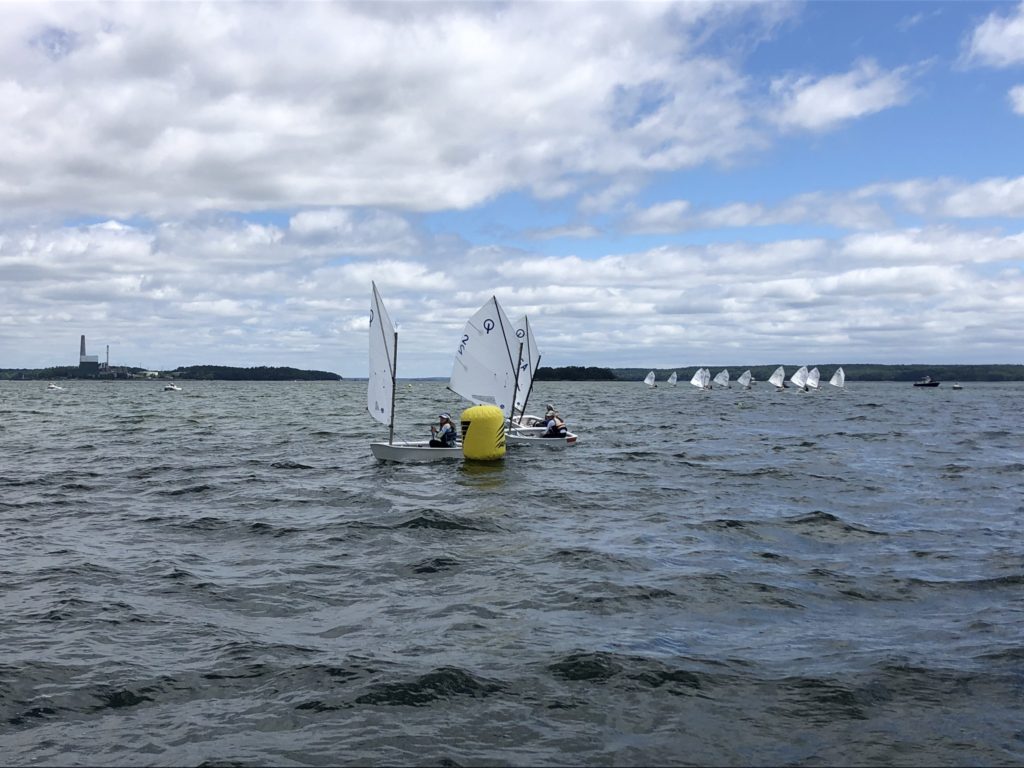 Small sailboats with white sails race around a yellow buoy on a choppy lake under a partly cloudy sky. More sailboats are visible in the distance, and land with a factory or plant is on the horizon.