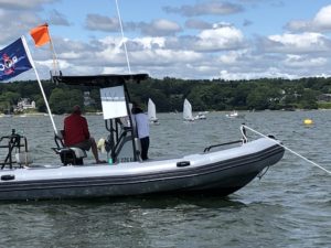 A rigid inflatable boat with two people aboard is anchored on a lake. Multiple small sailboats are visible in the background under a partly cloudy sky, with green trees and houses along the shoreline.
