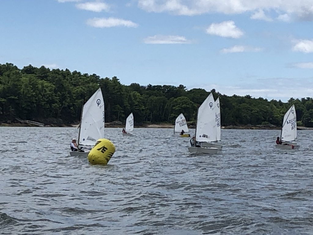 Several small sailboats with young sailors navigate around a yellow buoy on a lake, with a forested shoreline and a partly cloudy sky in the background.