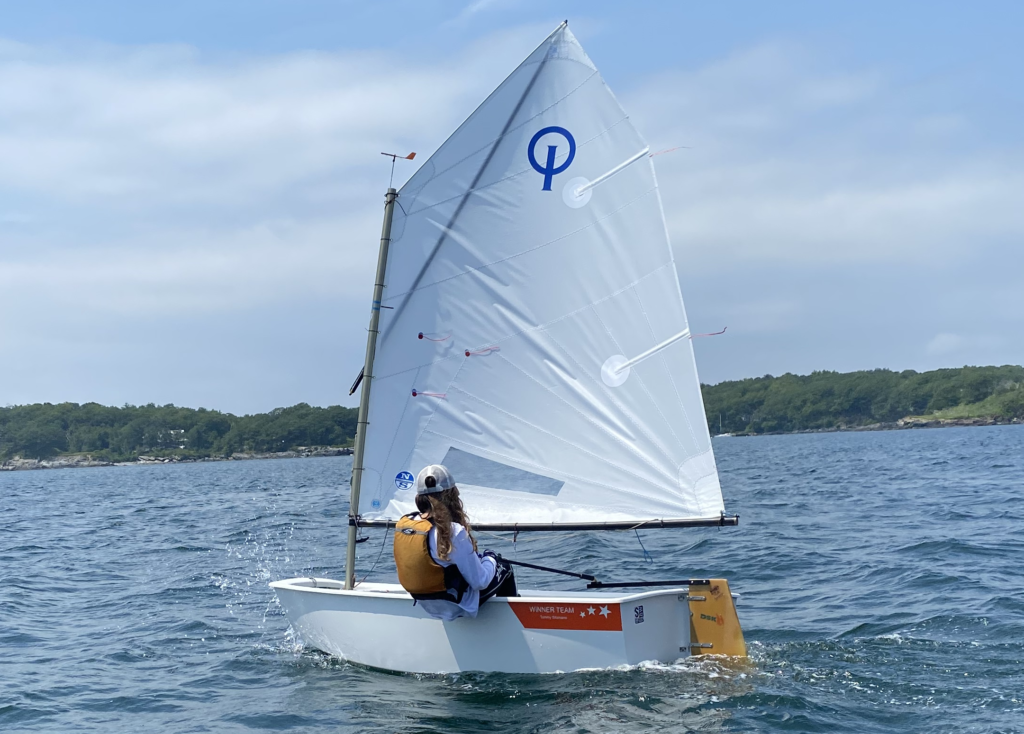 A person wearing a life jacket sails a small white dinghy with a single sail marked by a blue “Q” on calm water, with trees and a partly cloudy sky in the background.
