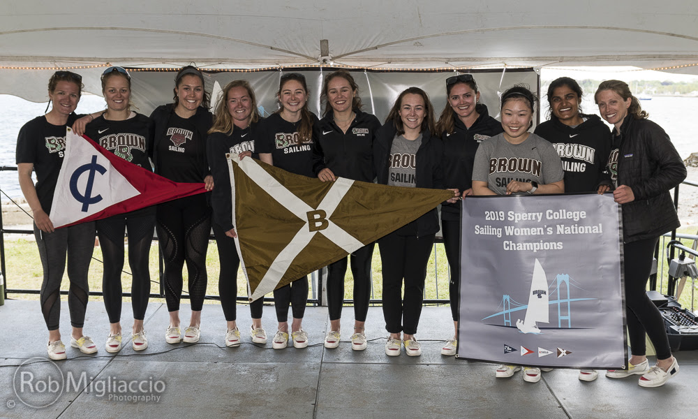 A group of women sailors from Brown University smile and stand together under a tent, holding sailing flags and a banner that reads '2019 Sperry College Sailing Women's National Champions.