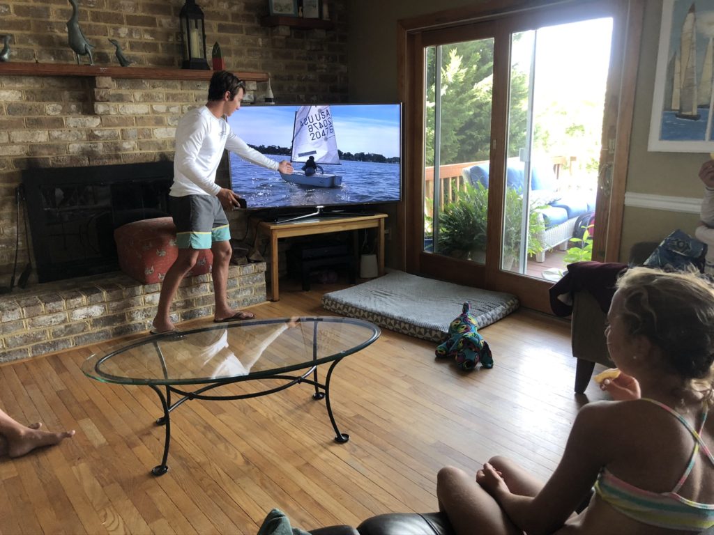 A man in shorts points at a sailboat shown on a TV screen in a living room, while two children watch. Sunlight comes in through a glass door, and a dog plush toy lies on the floor.