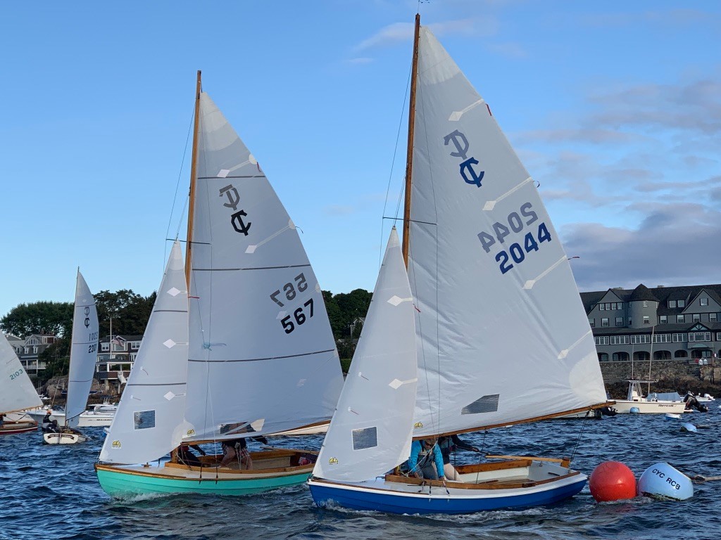 Two sailboats race side by side on the water near a red buoy, with people onboard. The sails display the numbers 567 and 2044. Houses and trees are visible along the shoreline under a blue sky.