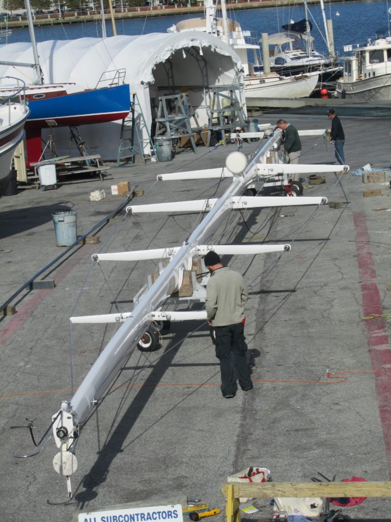 Several people work on a large sailboat mast laid horizontally on stands in a marina boatyard, with boats and covered structures visible in the background.