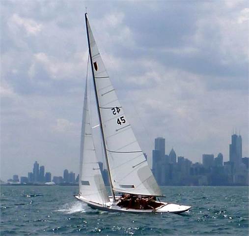 A white sailboat with several people on board sails on a choppy body of water, with a city skyline featuring tall buildings visible in the background under a partly cloudy sky.