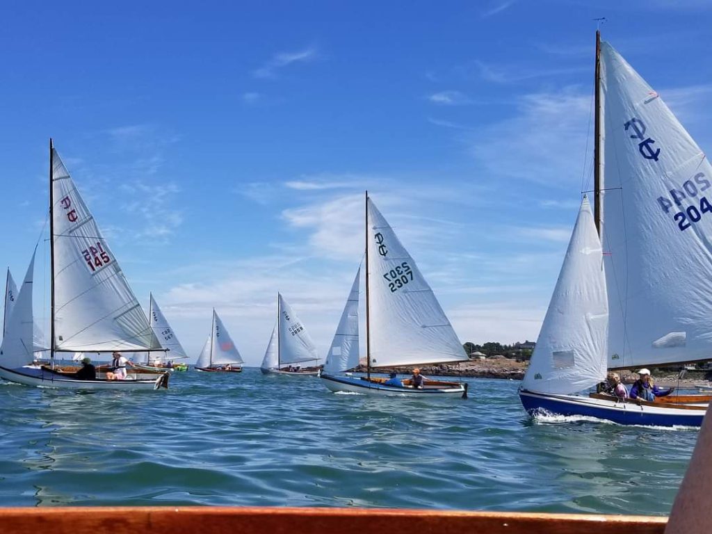 Several small sailboats with white sails are racing on a sunny day under a blue sky with scattered clouds. The water is calm, and people are visible on each boat, with a shoreline in the background.