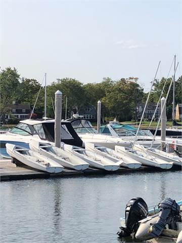 Several white motorboats are docked side by side at a marina, with a few sailboats and houses visible in the background. Trees and calm water surround the area under a clear sky.