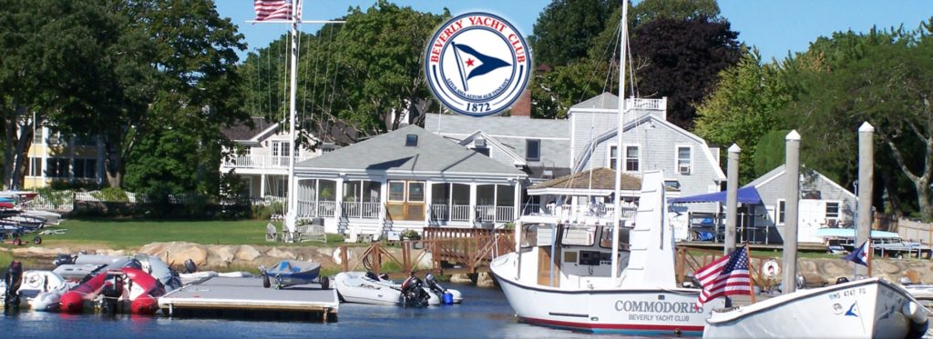 A marina with several boats docked near a white house and yacht club building. Trees and houses are in the background, with an American flag and a Beverly Yacht Club sign visible.