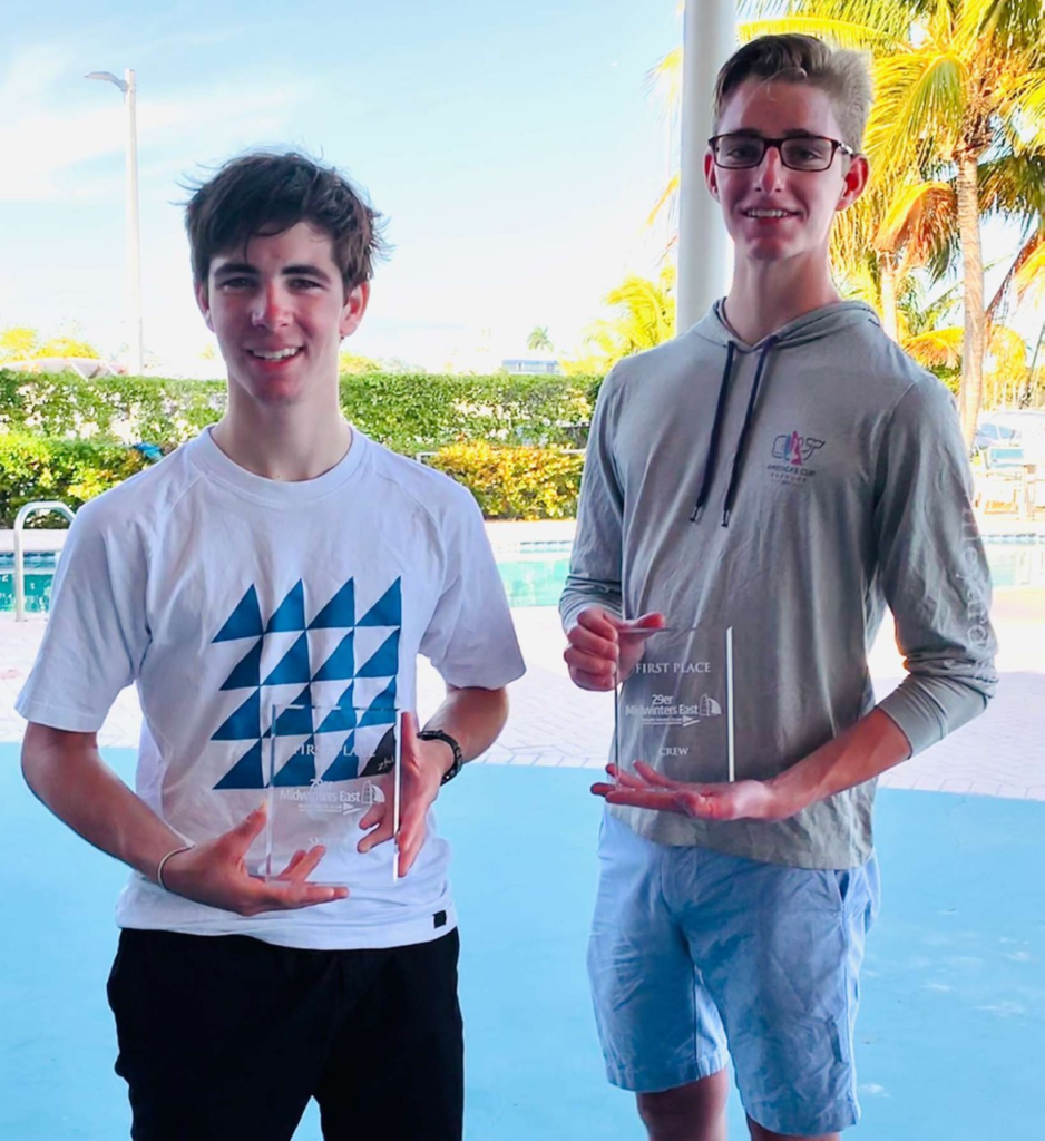 Two young men stand outdoors, smiling and holding clear glass trophies. Palm trees and a pool are in the background. One wears a white geometric t-shirt, the other a light hoodie and shorts. It appears to be a sunny day.