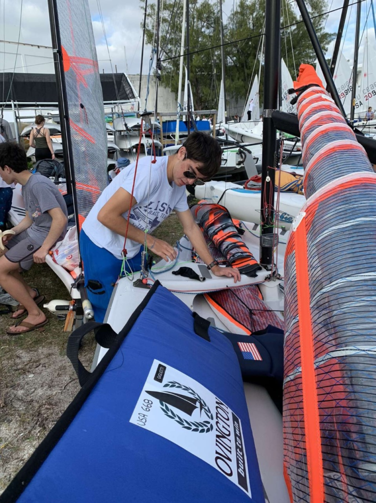 A person in sunglasses and a white t-shirt prepares a sailboat on land among other boats, with trees and a building in the background. Sail equipment and bags with championship logos are visible.