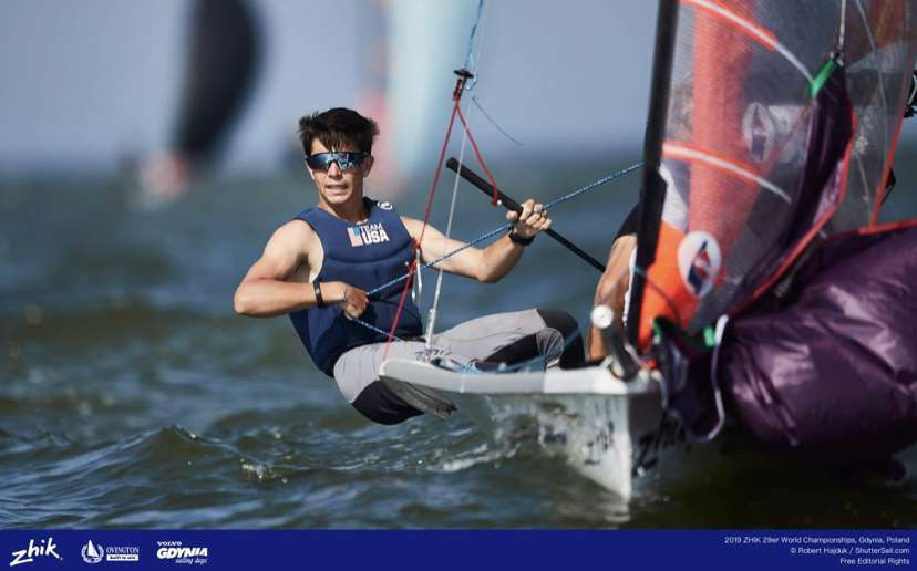 A male sailor wearing sunglasses and a navy tank top leans out of a small racing sailboat while holding a rope, navigating choppy water during a sailing competition. Other sailboats are visible in the blurred background.