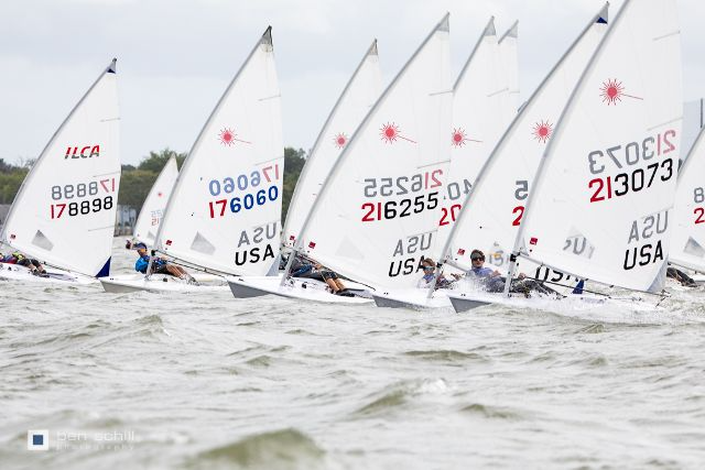 A group of Laser sailboats with sailors competing in a race, tilting on choppy water, with visible sail numbers and 'USA' markings. The sky is cloudy and land is in the background.