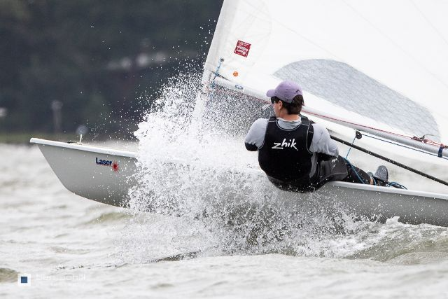 A sailor wearing a black vest and purple cap leans out of a small sailboat as water splashes up, with blurred trees and water in the background.