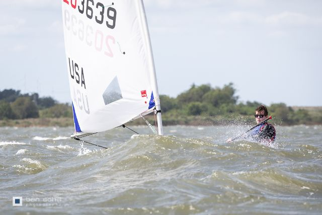 A person sailing alone in a small USA-marked sailboat on choppy water, partially obscured by a wave, with trees and a cloudy sky in the background.
