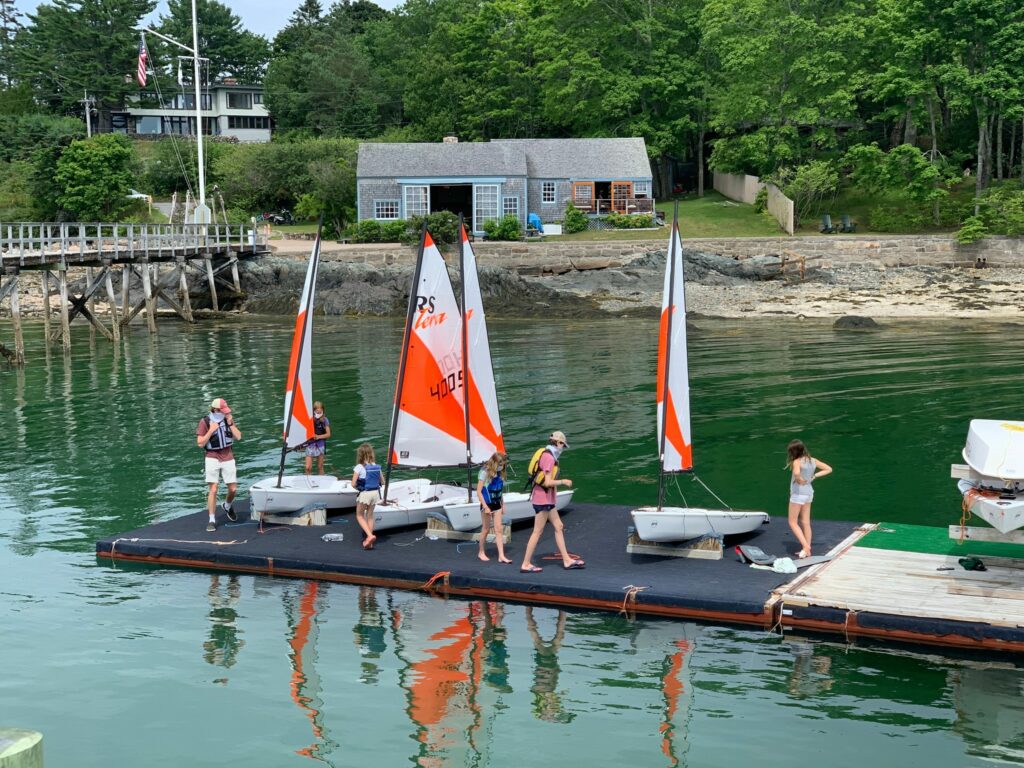 A group of people in life jackets prepare small sailboats on a floating dock by the shore, with a house, trees, and a wooden pier in the background. The water is calm and green.