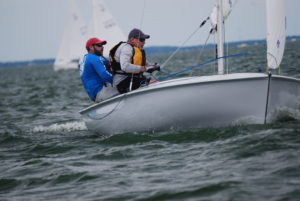 Two sailors wearing life jackets and caps are navigating a small sailboat on choppy water, leaning out for balance. Other sailboats are visible in the background under a cloudy sky.