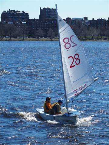 Two people are sailing a small white sailboat with the number 28 on its sail on a river, wearing life jackets. There are buildings and trees visible in the background under a clear blue sky.