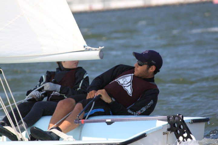 Two people wearing sunglasses, hats, and life jackets are sailing on a small boat. They are leaning out to balance against the wind, with water and a blurred shoreline in the background.